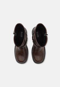 Botine din piele maro până la gleznă, cu o textură netedă, având un detaliu cu cataramă pe lateral, vârf rotunjit și cusături contrastante. Numele mărcilor "RAID" este vizibil în interior.