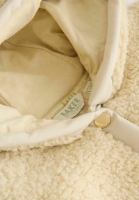 Kermanvärinen, pehmeältä tuntuva ulkokangas, jossa on beige vuori. Mukana kultaiset nappisuljenta ja näkyvät tarrat sisällä.