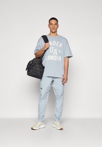 Camiseta oversized de color azul claro con el logo blanco de "Under Armour", combinada con joggers a juego de color azul claro, bolsa de hombro negra y zapatillas deportivas beige.