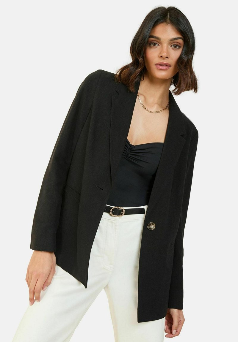 Černý blazer s přizpůsobeným střihem, zapínání na jeden knoflík a dvě kapsy. Nositý přes černé tričko a v kombinaci s bílými kalhotami.