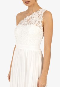 Bílé šaty s krajkovým designem na jednom rameni, přiléhavým živůtkem a splývavou sukní se zveřejněním, které zdůrazňuje jemnou texturu.