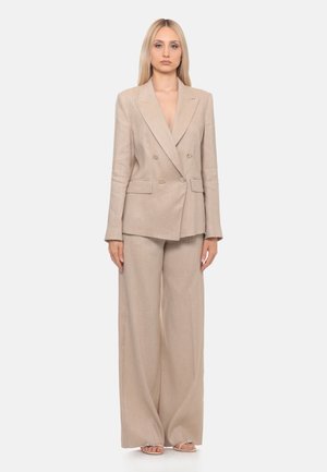 Donna bionda che indossa un blazer doppiopetto beige con pantaloni a gamba larga abbinati e sandali aperti, in piedi davanti a uno sfondo neutro.