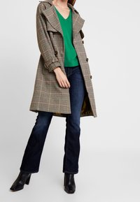 Hahnentritt-Trenchcoat mit großen Knöpfen, einem Reverskragen und Seitentaschen, kombiniert mit einem grünen V-Ausschnitt-Oberteil und dunkelblauen ausgestellten Jeans.