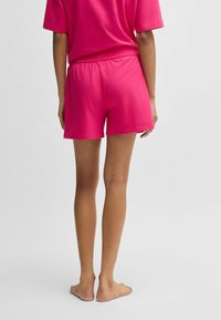 Fuchsia bomullsshorts med elastisk midja. Slät textur, rak snitt och längd till mitten av låret. Barfötter synliga.
