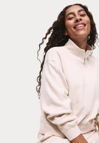 Beige fleece sweatshirt met een hoge kraag, verlaagde schouders en ribgebreide manchetten. Glad textuur, met een borstzakje.