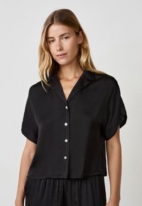 Blusa nera a maniche corte con una vestibilità rilassata, realizzata in tessuto liscio. Presenta un colletto e bottoni bianchi sulla parte anteriore.
