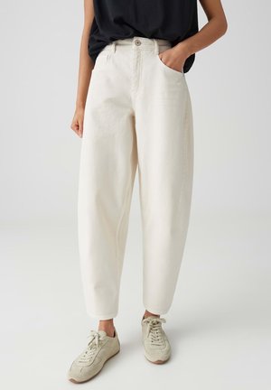 Person trägt weite, cremefarbene Jeans, beigefarbene Sneaker und ein schwarzes Oberteil, steht mit einer Hand in der Hosentasche vor neutralem Hintergrund.