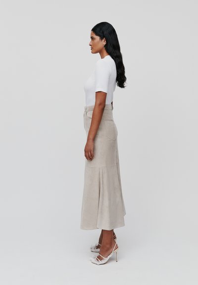 Weißes, eng anliegendes Oberteil kombiniert mit einem beigen Wildleder-Midi-Rock mit ausgestelltem Saum. Das Model trägt Riemchen-High Heels mit Federakzenten.