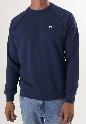 Homme portant un sweat-shirt bleu marine à col rond avec un petit logo ours blanc sur la poitrine gauche et un jean bleu clair.