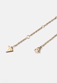 Kette aus vergoldetem Metall mit einem Karabinerverschluss und einem dreieckigen Anhänger, der mit "GUESS" und einem Fragezeichen graviert ist.