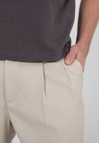 Homme portant un pantalon beige plissé et une chemise gris foncé, une main repose dans la poche droite du pantalon.