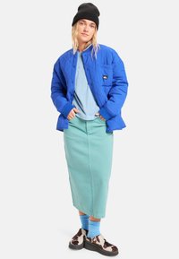 Modrá prošívaná bunda s kulatým límcem, doplněná světlomodrou košilí a mátově zelenou sukní. Hnědé a bílé vzorované boty a modré ponožky.