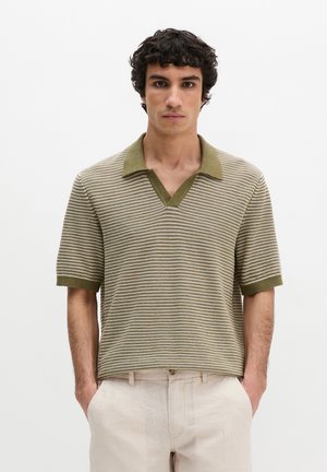 Mężczyzna o kręconych ciemnych włosach, ubrany w beżowo-oliwkową, prążkowaną, dzianą koszulkę polo z krótkim rękawem i jasnobeżowe spodnie, z rękami w kieszeniach.