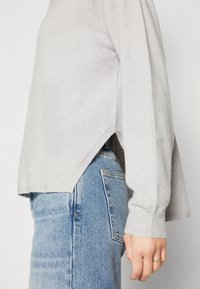 Vue latérale d'une personne portant un pull gris clair avec une fente sur le côté et un jean en denim bleu clair, la main reposant à côté de la hanche avec une bague en or visible.