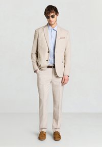 Beige suit with a light blue shirt and brown belt. Notable features include a brown pocket square, textured fabric, and sunglasses. Brown shoes.