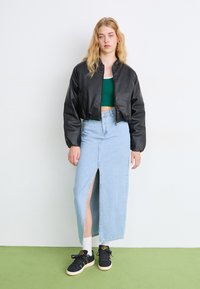 Μαύρο δερμάτινο μπόμπερ τζάκετ, πράσινη κοντή μπλούζα, ανοιχτό τζιν μαξι φούστα με πλαϊνό σκίσιμο, και μαύρα αθλητικά παπούτσια με χρυσές λεπτομέρειες.