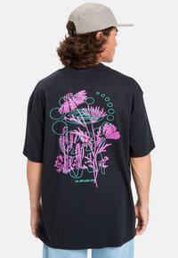Camiseta de algodón negra con mangas cortas, que presenta un diseño gráfico floral en rosa y turquesa en la parte posterior. Corte holgado, con escote redondo.