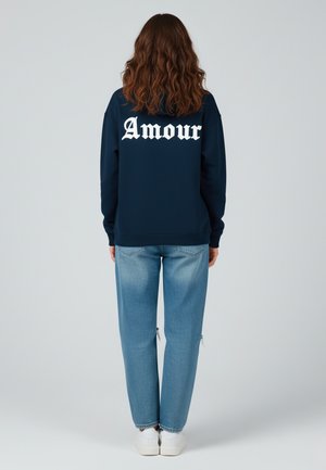 Felpa blu navy con testo bianco "Amour" sulla schiena, abbinata a jeans chiari strappati e sneakers bianche.