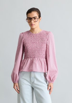 Γυναίκα με σκούρα μαλλιά και μεγάλα γυαλιά φοράει ένα ροζ, μακρυμάνικο, ριγέ πουκάμισο με σφηκοφωλιά στο μπούστο και ανοιχτό μπλε τζιν μπροστά σε γκρίζο φόντο.