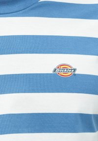 Μπλούζα με ριγέ μπλε και λευκές γραμμές από βαμβάκι, με κυκλικό λογότυπο Dickies σε κόκκινο, κίτρινο και μαύρο χρώμα στο μπροστινό μέρος.