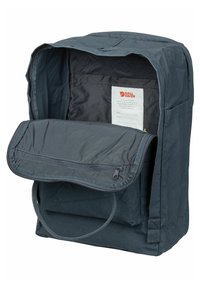 Zaino in tessuto grigio scuro con patta e tasca con zip. L'interno presenta un'etichetta e un rivestimento liscio. Spallacci resistenti e design minimalista.