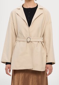 Beige Wickeljacke mit V-Ausschnitt und weiten Ärmeln, versehen mit einem taillierten Gürtel mit einem silbernen Ringakzent und glattem Stoff.