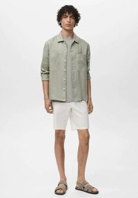 Svijetlozelena lanena košulja s kragnom, zatvaranjem na gumbe, dva prednja džepa, u kombinaciji s bijelim šortsevima i smeškim sandalama.