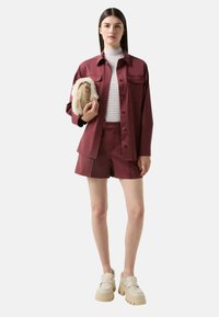 Burgunder satinjakke med brystlommer, hvidstribet turtleneck, matchende shorts og beige platform loafers. Holder en beige taske med pelsdetaljer.