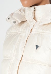 Lys beigefarget polstret jakke med høy krage, metallknapper og en trioangulært logo detalj på brystet. Myk, blank stofftekstur.