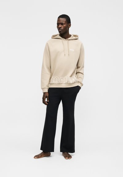 Homme portant un sweat à capuche beige avec un texte blanc et un pantalon large noir, debout pieds nus devant un fond blanc uni.