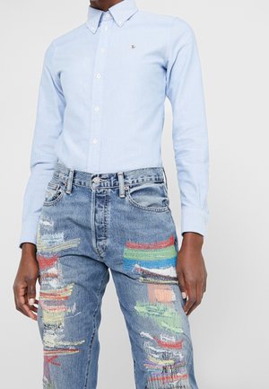 Chemise bleu clair à boutons associée à un jean bleu taille haute avec patchwork coloré et détails usés.