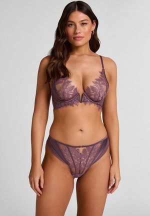 Woman with long brown hair wearing matching mauve lace bra and panties, standing against a plain light gray background.
