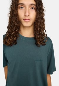 T-shirt in cotone color teal con collo rotondo classico e maniche corte, caratterizzato da un logo discreto sul petto. Tessuto morbido.