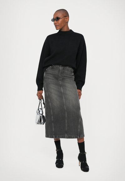 Pulover negru, fustă lungă din denim gri cu cusături vizibile, șosete negre, pantofi cu toc înalt și o poșetă argintie. Modelul stă în fața unui fundal alb.