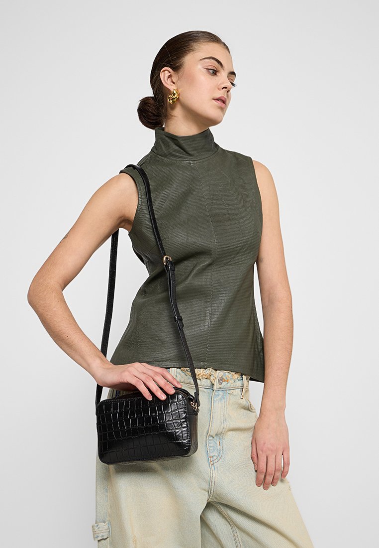 Grünes, ärmelloses Oberteil aus Leder mit hohem Kragen. Model hält eine kleine schwarze Handtasche mit Krokodilprägung. Hellblaue, weit geschnittene Jeans.