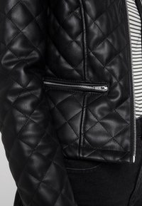 Veste en cuir rembourré noire avec une fermeture éclair diagonale et un tissu rayé en dessous. Présente une texture lisse et un motif losange distinct.