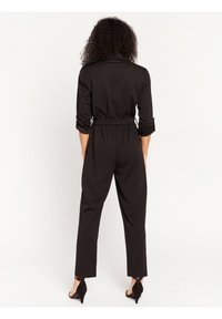 Black jumpsuit with long sleeves, elastic waistband, and tapered legs. Smooth fabric texture, minimalist design, and rolled sleeve details.