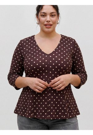 Mujer que lleva una camiseta marrón con cuello en V, lunares claros y mangas tres cuartos, con las manos juntas cerca de la cintura, de pie frente a un fondo liso.