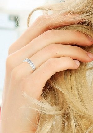 Bague en argent ornée d'une rangée de pierres précieuses rondes et claires, présentée sur une main tenant des cheveux longs et ondulés. Texture lisse, design simple.
