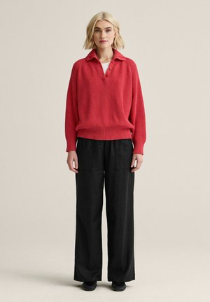 Kvinde iført en rød ribbet langærmet sweater og sorte bukser med vide ben, stående mod en ensfarvet beige baggrund.