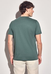 Zelené bavlněné tričko s kulatým výstřihem a krátkými rukávy, které vykazuje hladkou texturu a uvolněný střih, z pohledu zezadu.