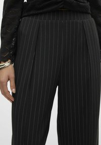 Pantalons noirs à rayures verticales avec une coupe ajustée, un devant plissé et un tissu texturé. Éléments matériels en argent simples sur la taille.