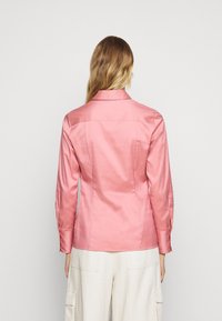 Camicia di raso rosa con maniche lunghe, colletto e polsini con bottoni. Texture liscia, vestibilità sartoriale e dettagli delle cuciture sul retro.