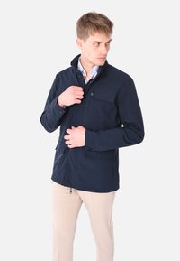 Giovane uomo che indossa una giacca blu navy sopra una camicia bianca e pantaloni beige, mentre aggiusta la cerniera guardando di lato.