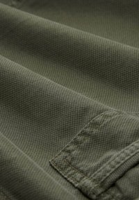 Tela verde oliva con una superficie texturizada, que presenta costuras dobladas y un detalle de bolsillo con parche. Costuras visibles en un color contrastante.