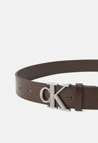 Ceinture en cuir marron avec une texture lisse, dotée d'une boucle en métal argenté en forme de "CK" et de trous régulièrement espacés pour l'ajustement.