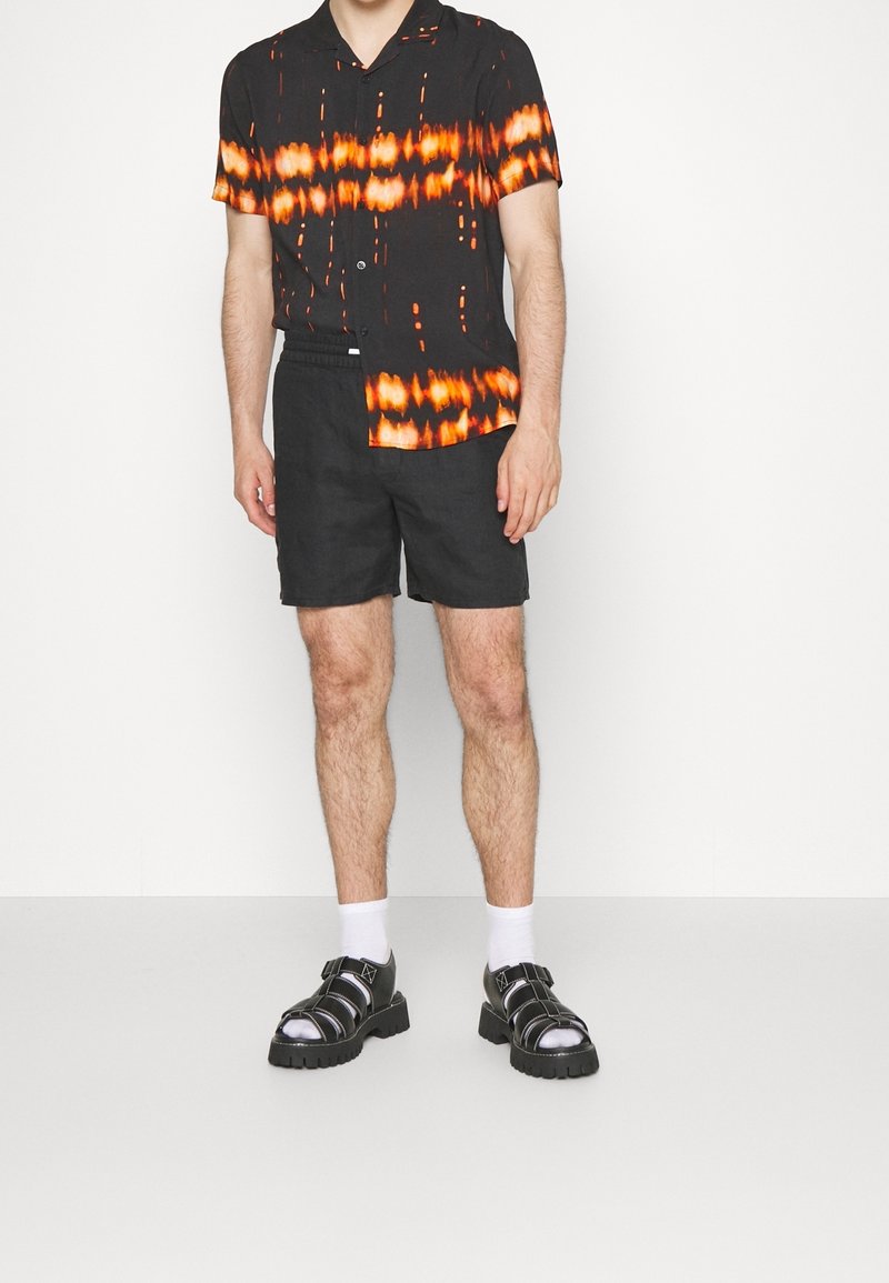 Zwart kortärmelig shirt met oranje en witte tie-dye patronen, gecombineerd met donkere korte broek en zwarte sandalen met zilveren accenten en dikke zolen.