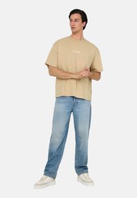 Beige T-Shirt mit kurzen Ärmeln und Rundhalsausschnitt sowie einem weißen Druck. Kombiniert mit hellblauen Jeans und weißen Sneakers.