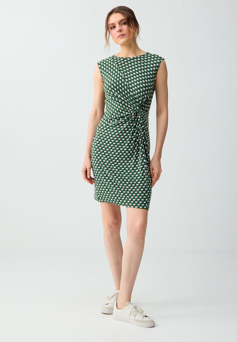 Femme debout portant une robe sans manches à motif géométrique vert et blanc avec un détail torsadé sur le côté et des baskets blanches sur fond uni.