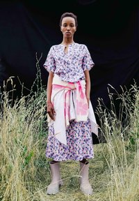 Bluse mit floralem Muster in Lila und Pink, kombiniert mit einem passenden Rock. Weißer Pullover um die Taille gebunden und graue kniehoge Stiefel.